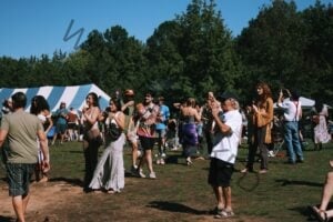 A lively crowd dances, claps, and mingles in a sunlit grassy field under a clear blue sky. People wear summer festival clothing, including tie-dye, crocheted tops, and flowing skirts. A blue-and-white striped tent stands at left and open-sided canopy tents are scattered behind, backed by a dense tree line.