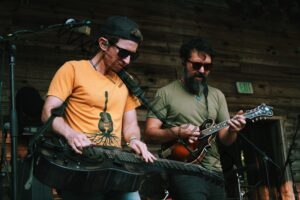 Two musicians perform side by side on a wooden stage. The man at left, in sunglasses, a backward cap, and an orange guitar-graphic T-shirt, plays a metal resonator guitar held flat in lap-steel style. Beside him, a bearded man in a green shirt and sunglasses plays a mandolin. Microphones and stage equipment surround them against a wooden back wall with a green "EXIT" sign.