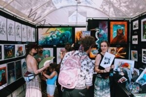 Inside a white festival tent, walls are covered floor to ceiling with framed artwork, including vivid paintings of herons and other animals and rows of black-and-white ink prints. Several people browse the booth, one carrying a pink backpack and another holding a green folding fan, while a woman at right smiles and holds up a black-and-white drawing for the camera.