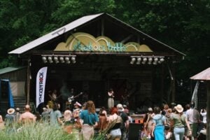A live band performs on the rustic wooden Shakori Hills stage, topped with a sign reading "shakori hills" in green and teal lettering beneath a peaked metal roof. A vocalist sings at the mic alongside guitarists, a bassist, and a drummer, with a white "School of Rock" banner at stage left. A large crowd in hats and summer clothes stands and watches from the grassy field, framed by tall green trees.