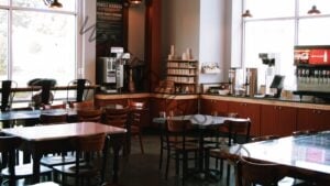 A warmly lit café interior with polished concrete floors and an assortment of wood tables and bentwood-style chairs. Along the back wall, a self-serve beverage station holds coffee brewers, stacked mugs on wooden shelving, and stainless iced-tea dispensers labeled "sweet" and "unsweet," set on light wood counters with rust-red cabinetry below. A large dark sign on the wall reads "The Root Cellar Family Dinners" and "Paleo Family Dinners," detailing the take-home meal program. Tall windows on either side let in daylight, and industrial pendant lights hang from the ceiling.