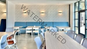 A café-style seating area inside 79° West featuring a long, channel-tufted blue banquette running along a wall papered in a soft, scalloped art-deco pattern and lit by three modern wall sconces. Small light wood tables sit in front of the banquette, paired with pale wood chairs. In the foreground, additional tables are surrounded by rolling chairs with gray mesh backs and rust-orange seats. Tall black-framed windows on the right look out onto a sidewalk and building exterior. The ceiling is dark and industrial with exposed ductwork and dome pendant lights.