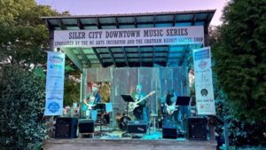 A band is performing at the outdoor concert venue at the NC Arts Incubator. A sign above the stage reads "Siler City Downtown Music Series". 