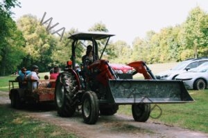 A man in sunglasses and a cap drives a red Massey Ferguson tractor with a front loader bucket, pulling a flatbed trailer loaded with straw bales. Several adults and children sit on the trailer for a hayride, with parked cars to the right and a tree line under a bright sky in the background.
