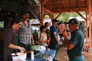 Under an open wooden pavilion strung with festoon lights, a woman serves food from a cast-iron skillet set on a portable burner at a table covered in a blue-and-white checked cloth, with stacked paper cups and a large green stockpot nearby. A man in a plaid shirt and cap stands beside her, and a line of people, including a child in a tie-dye shirt and an older man in a "Never Forget 9/11" cap, waits along the table. Trees, a rustic outbuilding, and parked cars are visible in the background.