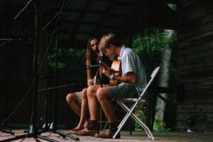 Two young men sit playing on a rustic wooden stage in low light, framed by several microphone stands. One in front, in a plaid short-sleeve shirt and shorts, leans over an acoustic guitar; the other, with long hair and a beard, plays a banjo beside him. Green trees and yellow wildflowers are visible through the open back of the stage.