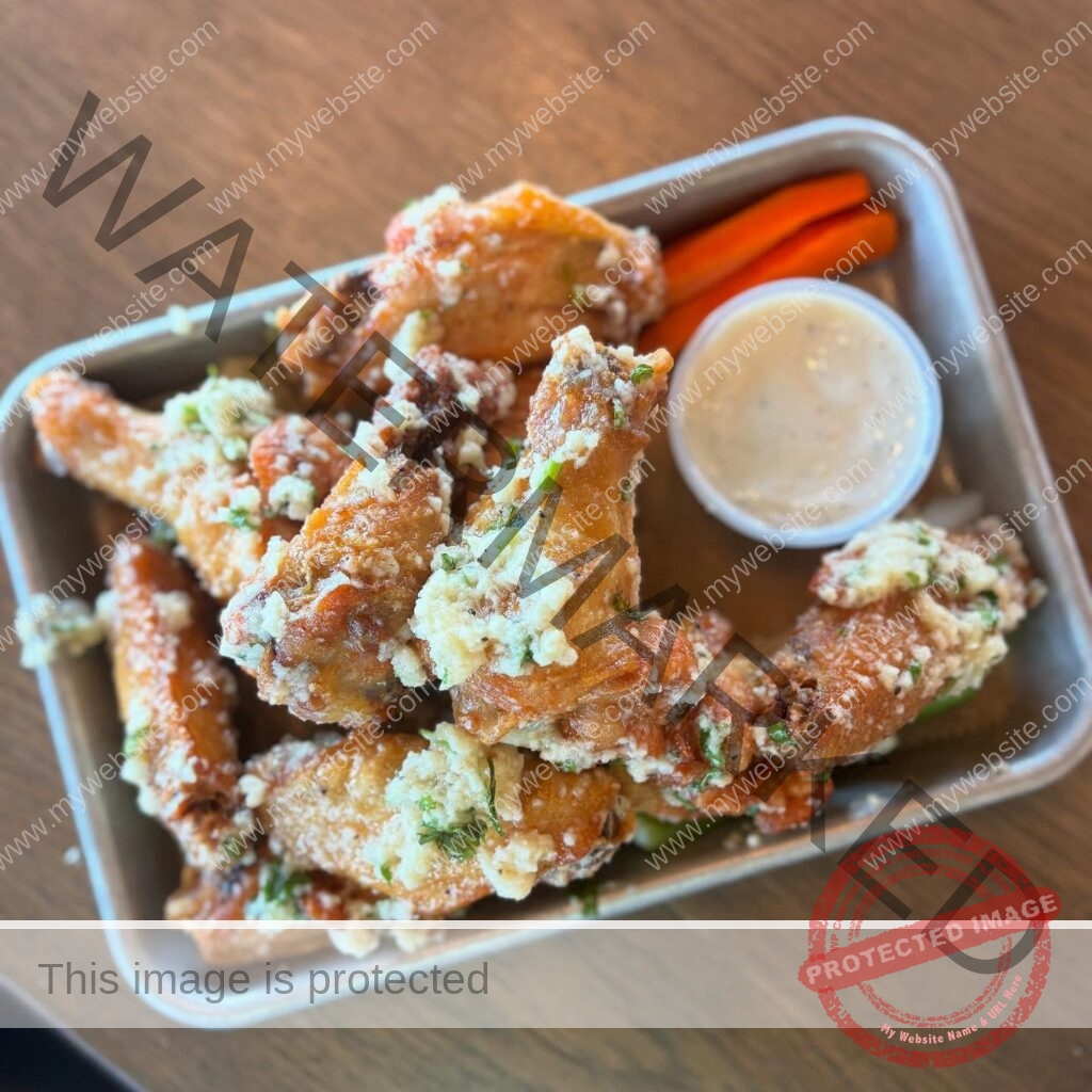 A close-up of a metal serving tin holding a generous portion of chicken wings at Hops & Berry, tossed in a garlic-parmesan coating and scattered with grated cheese and chopped fresh herbs. A small cup of creamy ranch or blue cheese dressing and a few carrot sticks sit alongside, set on a warm wood-toned table.