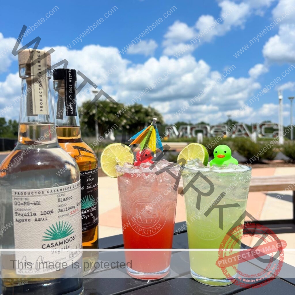 Two bright cocktails in branded Hops & Berry Taproom glasses sit on a black patio table under a blue, partly cloudy sky. The red drink and the green drink are each rimmed with salt, garnished with a lime wheel, and topped with a small rubber duck (one pink, one green) and a colorful paper cocktail umbrella. Two bottles of Casamigos tequila stand at left, and the large outdoor "MOSAIC" letter sculpture at Chatham Park is visible in the landscaped background.