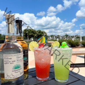 Two bright cocktails in branded Hops & Berry Taproom glasses sit on a black patio table under a blue, partly cloudy sky. The red drink and the green drink are each rimmed with salt, garnished with a lime wheel, and topped with a small rubber duck (one pink, one green) and a colorful paper cocktail umbrella. Two bottles of Casamigos tequila stand at left, and the large outdoor "MOSAIC" letter sculpture at Chatham Park is visible in the landscaped background.