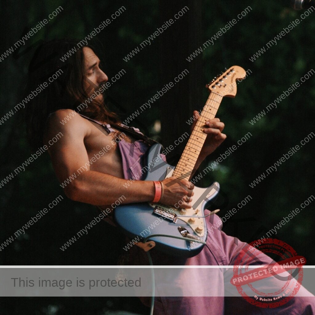 A man with long hair and a beard, shirtless under purple overalls, leans back as he plays a sky-blue Fender Stratocaster electric guitar. An orange festival wristband is on his wrist, and the background is dark with out-of-focus green foliage.