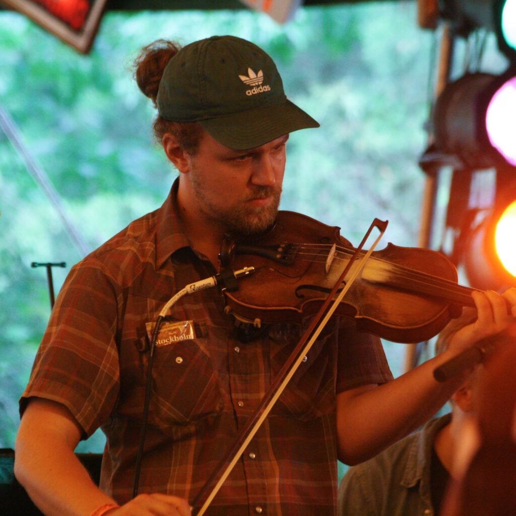 A man with a beard wearing a green Adidas cap and a brown plaid shirt plays a fiddle, drawing the bow across the strings with a clip-on mic attached to the instrument. Colored stage lights glow pink and white behind him, with green trees visible through the open side of the tent.