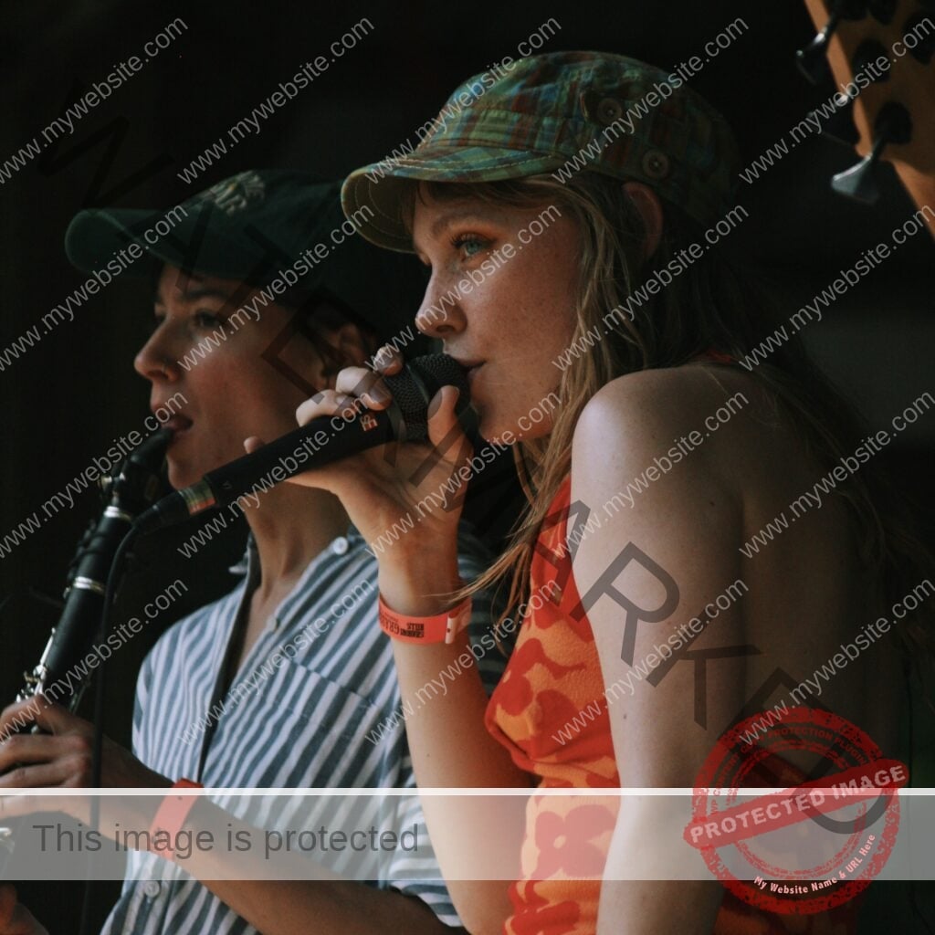 Two young performers sing side by side into a single microphone. The woman in front wears a green plaid cap and an orange floral halter top, with a Shakori Hills wristband on her wrist. Behind her, a person in a dark cap and striped shirt holds a clarinet. Warm side light falls across them against a dark stage background.
