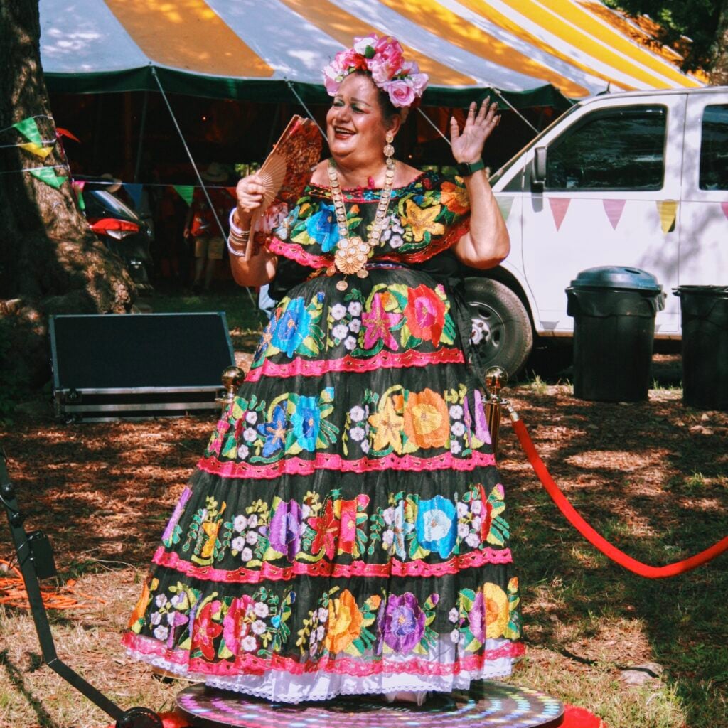 A performer stands on a small lighted platform wearing an elaborate black dress richly embroidered with multicolored flowers, a beaded necklace, and a headdress of pink roses. Smiling and mid-gesture, they hold a folded fan in one hand and raise the other. Behind them are a yellow-and-white striped tent, festival bunting, a white van, and trash bins on a wooded festival grounds.
