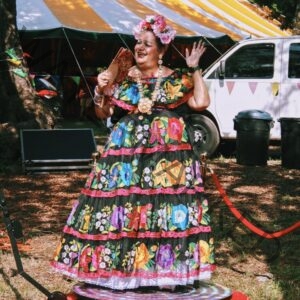 A performer stands on a small lighted platform wearing an elaborate black dress richly embroidered with multicolored flowers, a beaded necklace, and a headdress of pink roses. Smiling and mid-gesture, they hold a folded fan in one hand and raise the other. Behind them are a yellow-and-white striped tent, festival bunting, a white van, and trash bins on a wooded festival grounds.