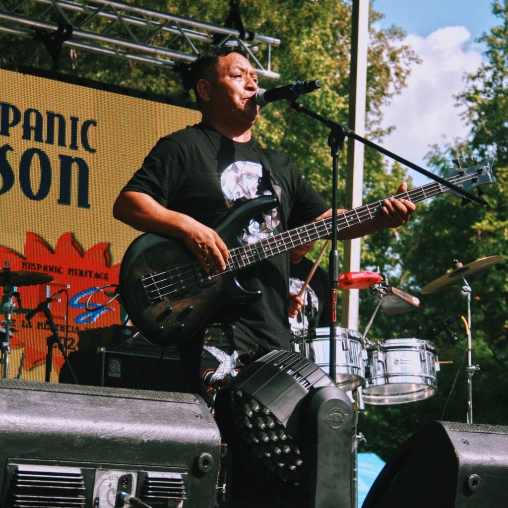 A man in a black graphic T-shirt sings into a microphone while playing a five-string electric bass on an outdoor stage. A large LED screen behind him reads "Hispanic Heritage," and a second musician plays timbales beside a drum kit. Stage trusses and green trees frame the scene under a partly cloudy sky.