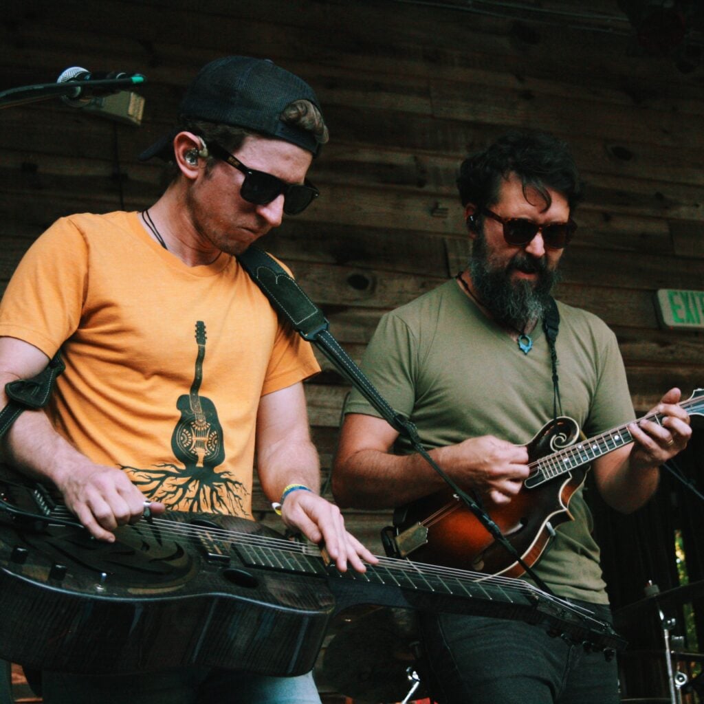 A close view of two musicians on a wooden stage. At left, a man in sunglasses, a backward cap, and an orange guitar-graphic T-shirt plays a metal resonator guitar held flat in lap-steel style. Beside him, a bearded man in sunglasses and an olive shirt plays a mandolin. A green "EXIT" sign and stage equipment are visible against the wooden back wall.