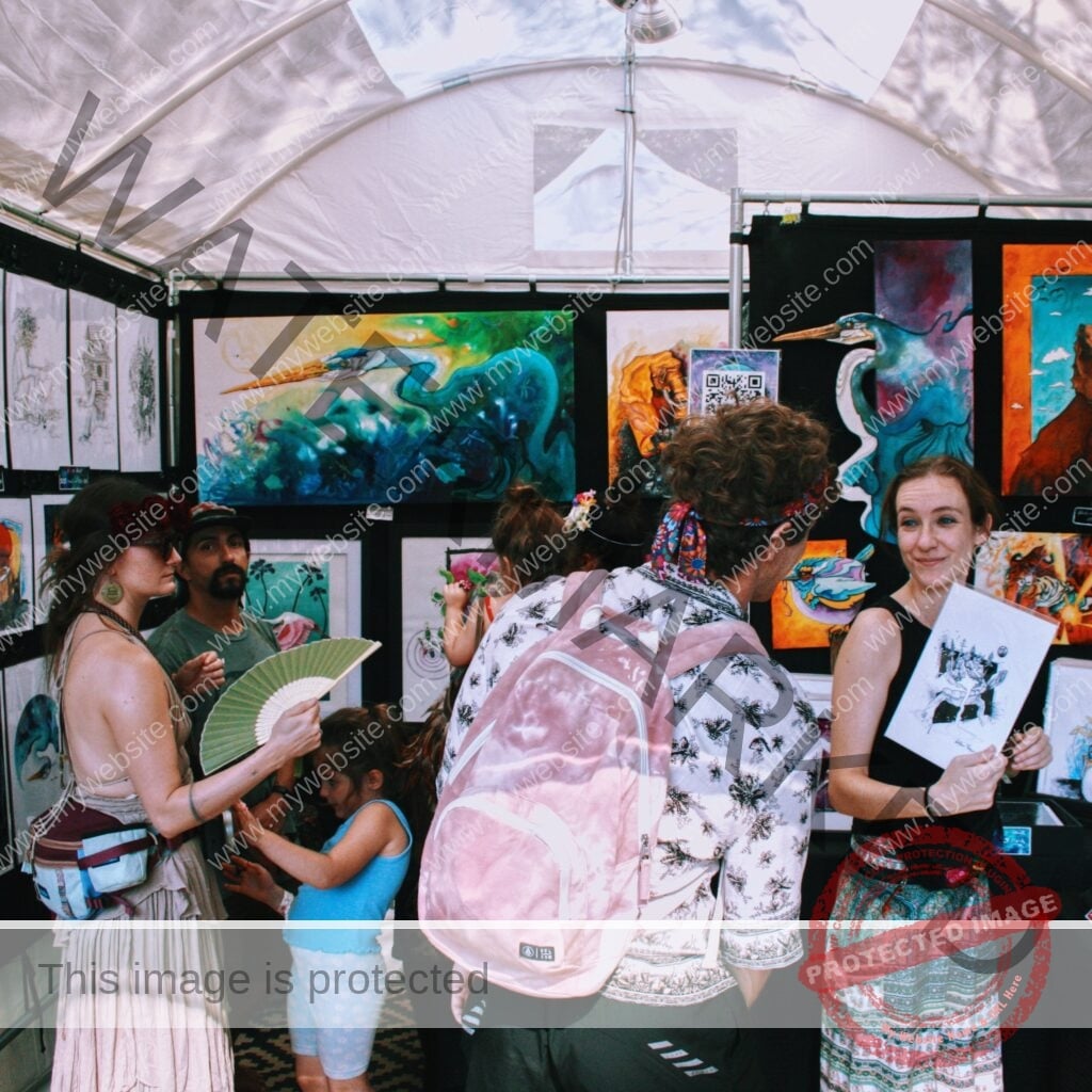Inside a white festival tent, the walls are covered with framed artwork, including vivid paintings of herons and other animals and rows of black-and-white ink prints. Several people browse the booth, one carrying a pink backpack and another holding a green folding fan, while a woman at right smiles and holds up a black-and-white drawing toward the camera.