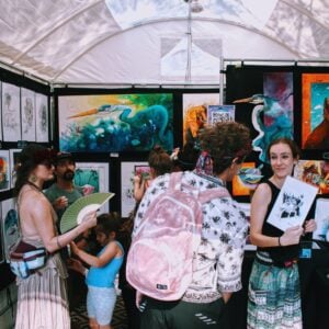Inside a white festival tent, the walls are covered with framed artwork, including vivid paintings of herons and other animals and rows of black-and-white ink prints. Several people browse the booth, one carrying a pink backpack and another holding a green folding fan, while a woman at right smiles and holds up a black-and-white drawing toward the camera.