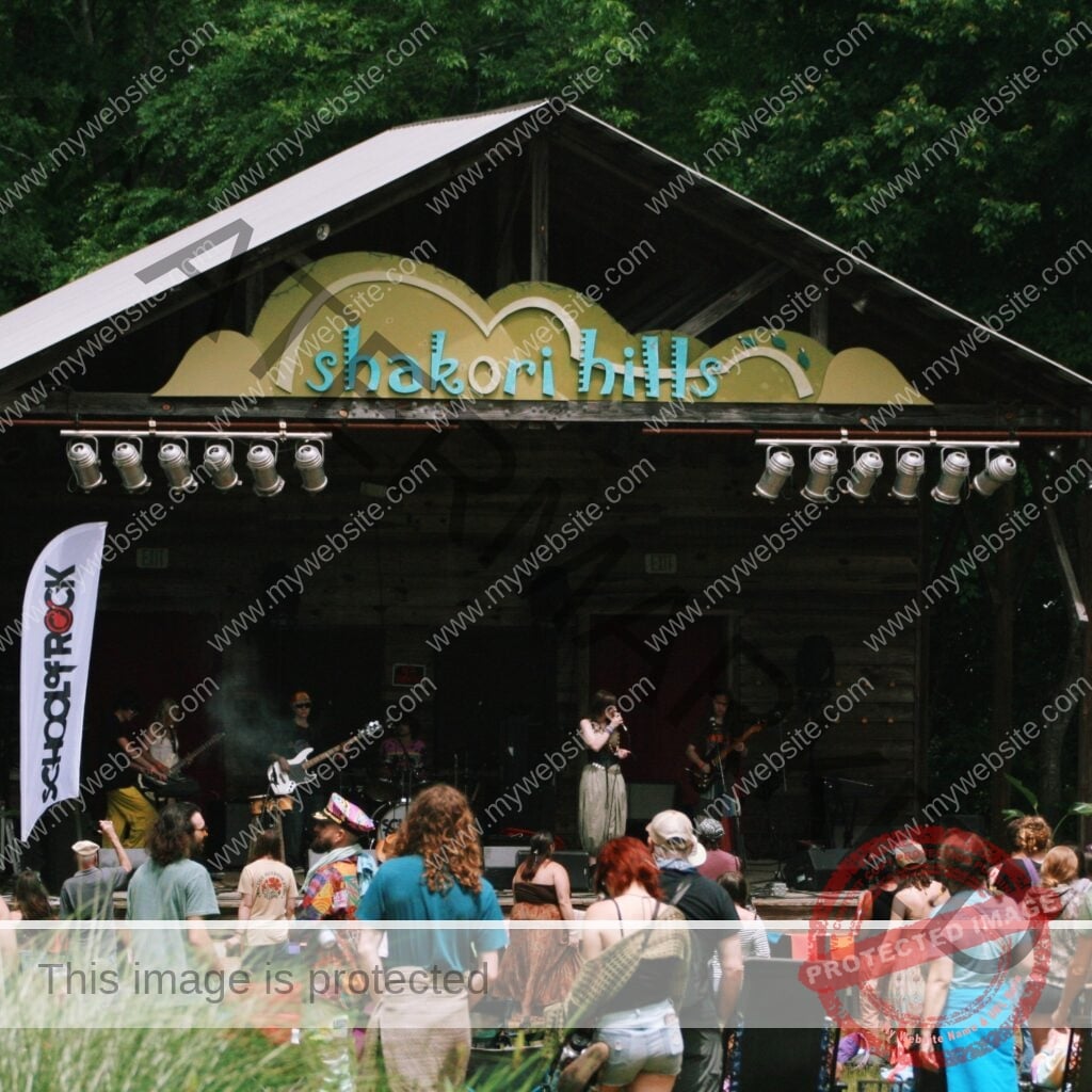 A live band performs on the rustic wooden Shakori Hills stage, topped with a green-and-teal "shakori hills" sign under a peaked metal roof. A vocalist sings at the mic alongside guitarists, a bassist, and a drummer, with a white "School of Rock" banner at stage left. A crowd in hats and summer clothing watches from the grassy field in front.