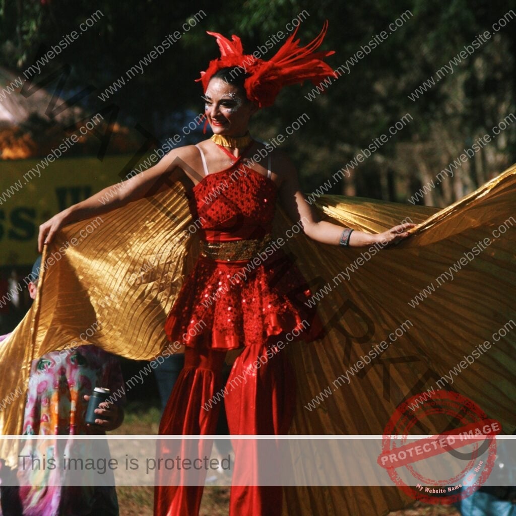 A performer on stilts wears a red sequined costume with flared red pants, a gold sequined waist sash, a feathered red headdress, and face glitter. They spread a pair of large gold fabric wings wide and smile. A festivalgoer in a tie-dye outfit holds a drink at lower left, with trees and a banner in the background.