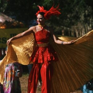 A performer on stilts wears a red sequined costume with flared red pants, a gold sequined waist sash, a feathered red headdress, and face glitter. They spread a pair of large gold fabric wings wide and smile. A festivalgoer in a tie-dye outfit holds a drink at lower left, with trees and a banner in the background.
