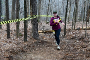 A woman runs along a packed-dirt trail through leafless winter woods, carrying a yellow relay baton. She wears a maroon long-sleeve shirt with a graphic print, black leggings, an olive cap, and a yellow race bib reading "RELAY 383." The course is lined with black-and-yellow striped caution tape strung between the trees, and the ground is covered in brown fallen leaves and pine needles.