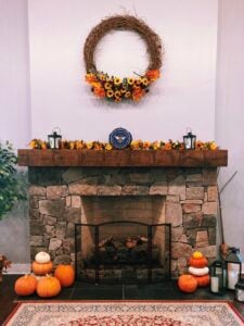 A large stacked-stone fireplace with a thick rustic wood mantel and a black metal screen across the hearth. The mantel is decorated with a sunflower garland, two black lanterns, and a round bee-motif sign; a grapevine wreath trimmed with sunflowers and autumn leaves hangs on the white wall above. Clusters of orange and white pumpkins, candles, and lanterns sit on the slate hearth, with a patterned area rug in the foreground and a leafy plant to the left.