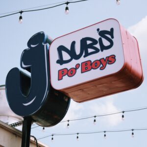 J Dub's Po'Boys sign with string lights in the background against a cleary blue sky. This new eatery will provide food for the Sake Tutored Tasting.