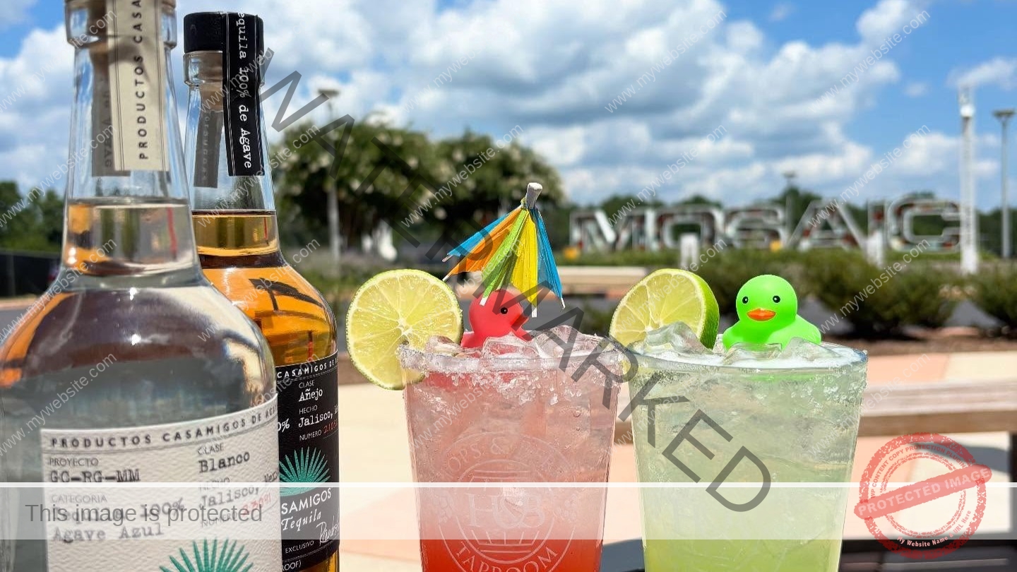 Two bright cocktails in branded Hops & Berry Taproom glasses sit on a black patio table under a blue, partly cloudy sky. The red drink and the green drink are each rimmed with salt, garnished with a lime wheel, and topped with a small rubber duck (one pink, one green) and a colorful paper cocktail umbrella. Two bottles of Casamigos tequila stand at left, and the large outdoor "MOSAIC" letter sculpture at Chatham Park is visible in the landscaped background.
