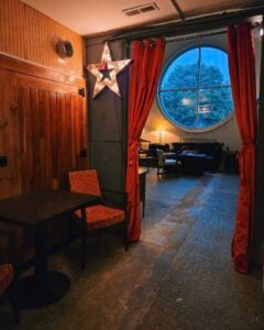 A cozy, dimly lit interior with a wood-paneled wall and a black café table with a floral-upholstered chair on the left. A large star fixture studded with glowing bulbs hangs on the wall, and tied-back red velvet curtains frame an opening into a lounge area with sofas, a lamp, and a tall round window looking out onto trees at dusk.