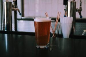 A pint glass of amber beer topped with a white foam head sits on a dark countertop. Behind it, stainless-steel tap towers, a cup of straws, and white subway tile fill the softly blurred background of a brewery taproom.