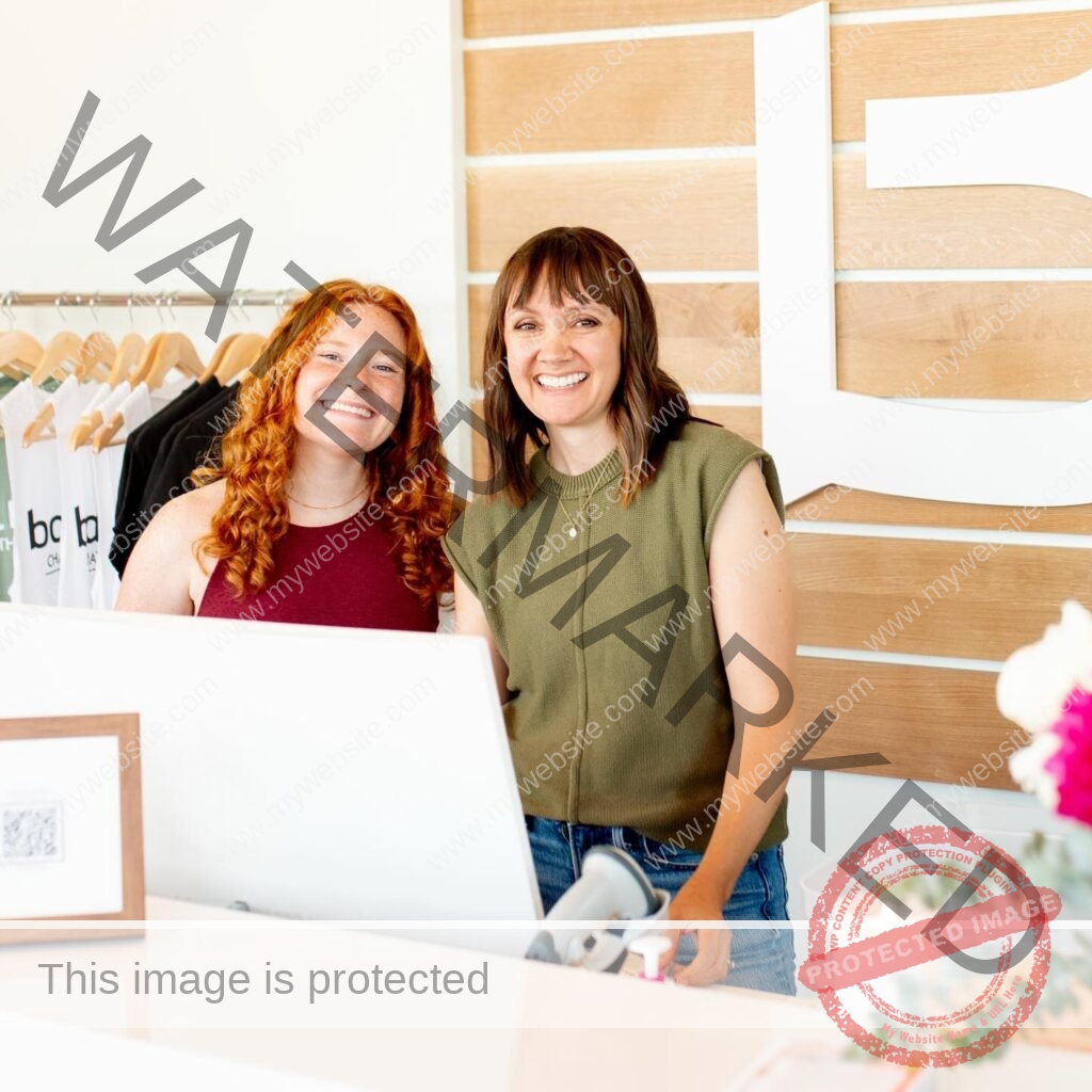 Two women stand smiling behind a white reception desk inside the barre3 Chatham Park studio. The woman on the left has long, curly red hair and wears a maroon tank top; the woman on the right has dark shoulder-length hair with bangs and wears an olive-green sleeveless top with jeans. Behind them, a rack holds light-colored branded apparel, and a wall of pale wood slats displays a large white "b3" logo. The counter in the foreground holds a white laptop, a small framed sketch, a barcode scanner, and a potted plant with pink blooms at the right edge.