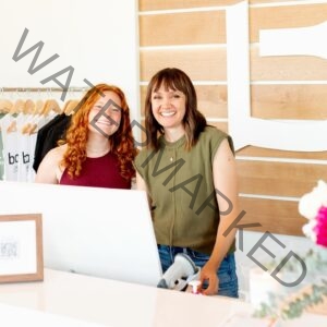 Two women stand smiling behind a white reception desk inside the barre3 Chatham Park studio. The woman on the left has long, curly red hair and wears a maroon tank top; the woman on the right has dark shoulder-length hair with bangs and wears an olive-green sleeveless top with jeans. Behind them, a rack holds light-colored branded apparel, and a wall of pale wood slats displays a large white "b3" logo. The counter in the foreground holds a white laptop, a small framed sketch, a barcode scanner, and a potted plant with pink blooms at the right edge.