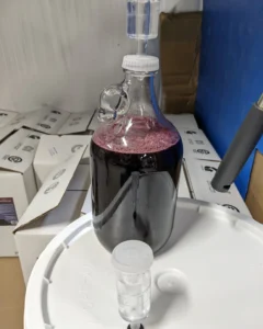 A glass growler-style jug filled with deep purple-red fermenting fruit mead, topped with a drilled cap and a clear plastic airlock. A pinkish layer of fermentation foam sits on the surface of the liquid. The jug rests on a white food-grade bucket lid, with a second airlock in the foreground and stacked white shipping boxes behind it in the production area.