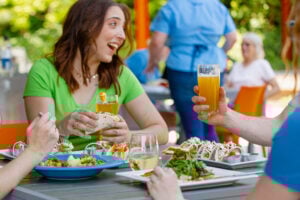 A woman in a green t-shirt laughs mid-conversation at an outdoor restaurant table, holding a taco and a glass of beer. The table is filled with plates of tacos, salad, and a blue bowl of roasted vegetables, with glasses of beer and white wine. Other diners and a server in a blue apron are visible in the background among lush green trees.