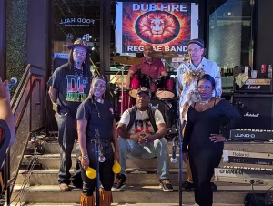 Six members of the Dub Fire Reggae Band posing on stage in front of their backdrop banner, which features a fiery lion's head logo. The group stands around a drum kit and Roland and Yamaha keyboards, with Marshall amplifiers and a "Food Hall" sign visible behind them. Several members wear "DF" band t-shirts.