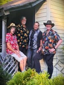 Four men pose together on a porch in front of a clapboard building, all smiling at the camera. They wear colorful Hawaiian and floral-print shirts; two wear hats, and one sits on a porch rail. Green shrubs frame the bottom of the frame.