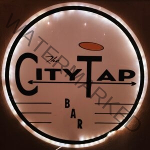 Circular backlit sign for "The City Tap Bar" mounted on a dark wall. The logo features black mid-century-style lettering on a white face, with an orange oval floating above the "T" and horizontal lines flanking the word "Bar" at the bottom. A ring of white LED light illuminates the sign from behind.