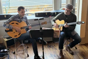 Two acoustic guitarists perform together inside a sunlit storefront venue with large windows and a warm wood-plank floor. On the left, a man in a blue plaid flannel shirt plays a blonde archtop guitar with distinctive oval f-holes; on the right, a man in a flat cap, glasses, dark zip pullover, and brown boots plays a classical-style acoustic guitar. A shared music stand with sheet music sits between them, with a small AER amplifier, instrument cases, and tangled cables visible around their feet. Outside the windows, daylight illuminates a quiet street and curb beyond.