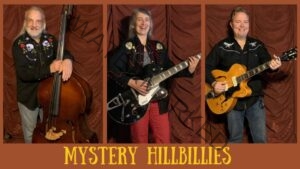 Promotional band photo for the Mystery Hillbillies, a trio shown in three side-by-side portraits against a rust-red curtain. Left: a bearded man in an embroidered western shirt holding an upright bass. Center: a smiling woman in a western shirt and red pants playing a black hollow-body electric guitar. Right: a man in a western shirt playing a blonde hollow-body electric guitar. "Mystery Hillbillies" is set in yellow western-style lettering across the bottom.
