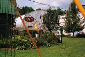 The exterior of Fair Game Beverage Co. at The Plant in Pittsboro, featuring a distinctive silver Quonset hut-style building with the Fair Game Beverage Co. oval sign, a wooden deck with a yellow umbrella, string lights overhead, lush green landscaping, and a chalkboard sandwich sign in the foreground.