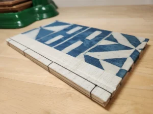 A handmade stab-bound book resting on a light wood surface, viewed from the spine edge. The cover is wrapped in cream and indigo-blue fabric printed with a geometric pinwheel-and-bar pattern, and the spine shows the exposed dark thread of Japanese-style stab binding stitched through the pages. A green object sits blurred in the background.