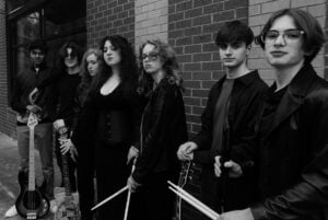Black-and-white band portrait of seven young musicians posing against a brick wall, all dressed in black with serious expressions. Two hold electric bass guitars at their sides, one holds a microphone stand, two hold drumsticks, and one holds a violin bow. The styling reads as moody alt-rock or goth-leaning.