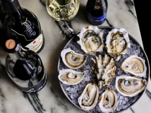 Overhead view of a dozen fresh oysters on the half shell served on crushed ice, arranged on a marble surface alongside two wine bottles and a poured glass of white wine.