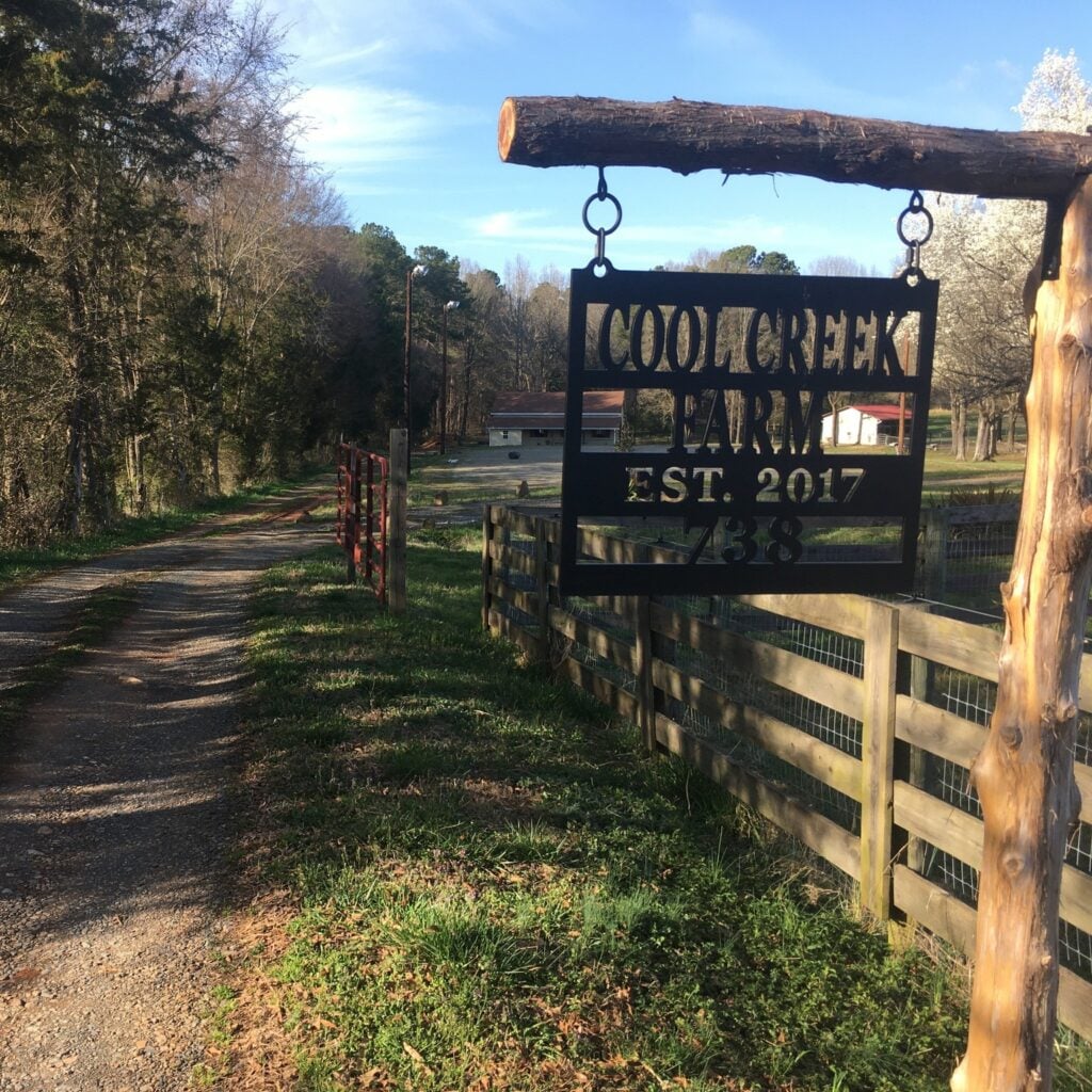 The entrance to Cool Creek Farm, with a black metal hanging sign suspended from a rough-hewn cedar log crossbeam reading "COOL CREEK FARM EST. 2017 738." A gravel driveway curves to the left toward a barn and farm buildings in the background, lined by a wooden split-rail fence with wire backing on the right and bare early-spring trees on the left. White flowering trees bloom in the distance under a clear blue sky.