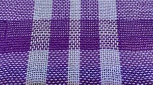 Close-up of a handwoven textile showing an intricate plaid or windowpane pattern in deep purple and pale lavender threads, with the individual warp and weft threads clearly visible.