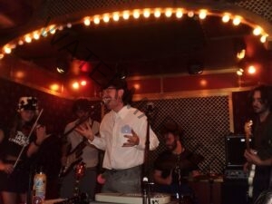  Live performance photo of a five-piece band on a small stage framed by a curved arch of warm-toned marquee lights. A lead vocalist in a white button-down shirt sings at the center microphone with an expressive gesture; a violinist in a brimmed hat performs on the left, alongside another guitarist, a drummer behind the kit, and a bass guitarist on the right. The stage has a red-and-cream checkered back wall, giving the space an intimate honky-tonk or roadhouse atmosphere.