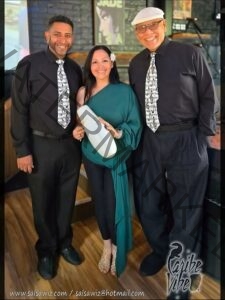 Promotional photo of the trio Caribe Vibe posed indoors on a wooden floor. Two men in black shirts and musical-note-patterned ties flank a woman in a flowing teal outfit with a white flower in her hair; she holds a white güiro (a Latin percussion instrument). The Caribe Vibe logo appears in the lower right, with the website salsawiz.com and email salsawiz@hotmail.com along the bottom.