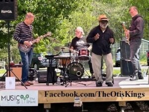 The Otis Kings performing live on an outdoor stage, with a guitarist in a striped shirt on the left, a drummer behind a kit branded with "The Otis Kings" and a crown logo, a lead vocalist in a fedora and dark shirt at center, and a bassist on the right. A "Music @ The Plant" sign is visible on the stage front alongside the URL Facebook.com/TheOtisKings.