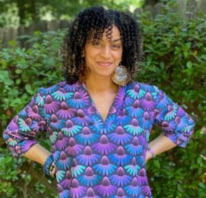 Outdoor portrait of a woman with shoulder-length curly dark brown hair with lighter highlights, smiling warmly at the camera with her hands on her hips. She wears a v-neck tunic blouse with a bold botanical print of purple, blue, teal, and pink coneflowers on a dark gray background, paired with large statement earrings that appear to be made of natural wood and stone or shell. Behind her is a lush green garden hedge and a weathered wooden fence, with leafy trees visible in the background.